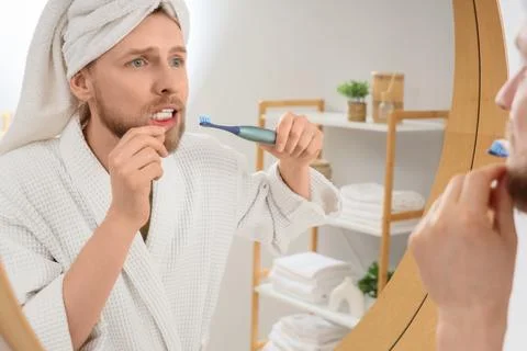 Young man brushing teeth with electric toothbrush near mirror in bathroom Stock Photos