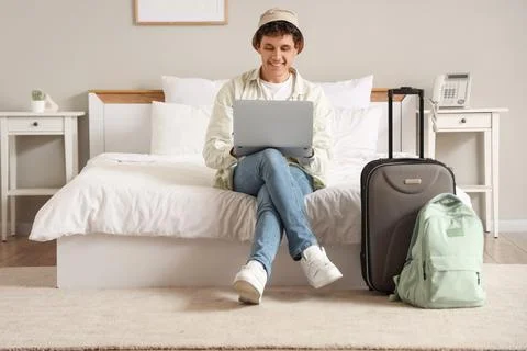 Young man in a bucket hat using a laptop on a hotel bed with luggage, remote  Stock Photos