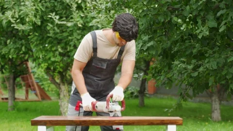 Young man builder carpenter grinds a wooden table with a grinding machine in the Vídeos de archivo 219998749