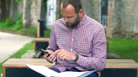 Young man calculating bills on smartphone sitting on in city park Vídeo Stock 100476319