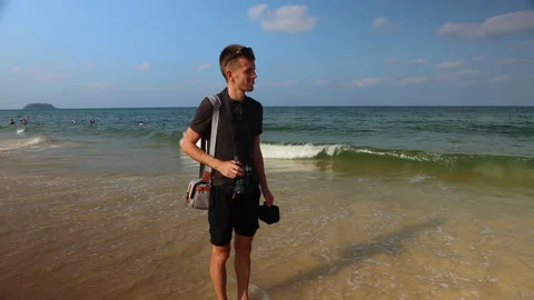 Young man with a camera stands on the beach by breaking waves. 스톡 동영상 146012960