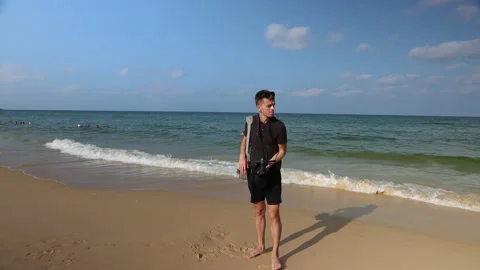 Young man with a camera stands on the beach by breaking waves. Sea background. 스톡 동영상 146072249