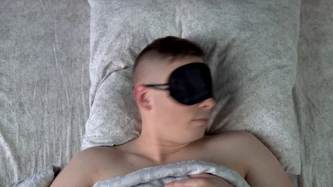 A young man cannot fall asleep in a sleep mask. A man lies in bed in his room Stock Footage 128015344