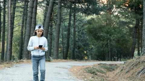 A young man in a cap and jeans walking along a forest path using the cell phone Stock Footage 141339142