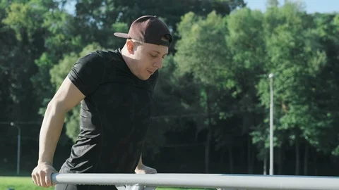 Young man in cap doing triceps dips on parallel bars at sunny morning. Side view 動画素材 115335071