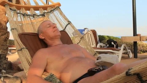 A young man in a cap from the sun is resting in a hammock on the seashore on a s Vidéo 115664200