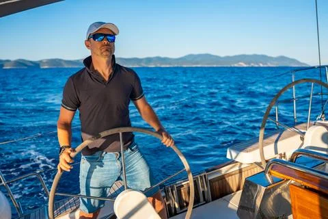 Young man captain stands at the helm and controls a sailboat during a journey by Stock Photos