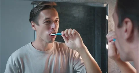 Young man carefully brushes teeth in bathroom in front mirror, observing rules Stock Footage 267873276