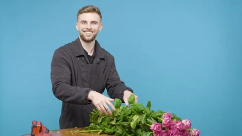 Young man carefully stacks pink roses on the wooden table on blue background Stock Footage 276378774