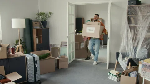 Young Man Carrying Two Moving Boxes and His Girlfriend with Plant Entering Room Stock Footage 323299396