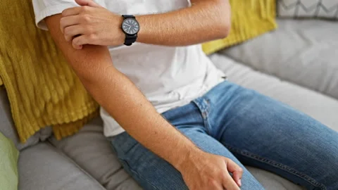 A young man in casual attire scratching his arm while sitting on a sofa ind.. 스톡 동영상 273003229