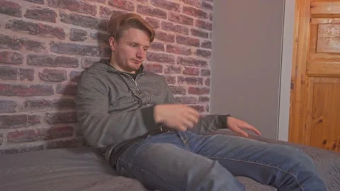 Young man casually sits down on the bed, rubs his hair, and starts using Stock Footage 212234596