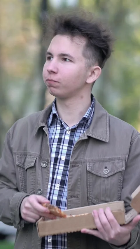 Young man casually walks down the street outdoors eating a slice of pizza from a Video stock 348790171