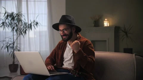 Young man celebration success using laptop siting at couch.Bearded guy wear hat Stock Footage 212316356