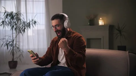 Young man celebration success using smartphone siting at couch.Bearded guy wear Stock Footage 212316542
