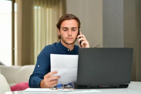 Young man checking a bill while using his phone in front of a laptop on an ou Stock Photos