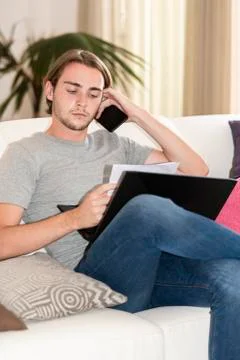 Young man checking some notes, holding his phone and a laptop while sitting o Stock Photos