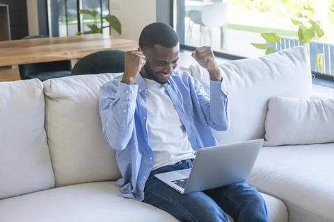 Young man cheers while using a laptop on a bright living room couch, expres.. Foto stock