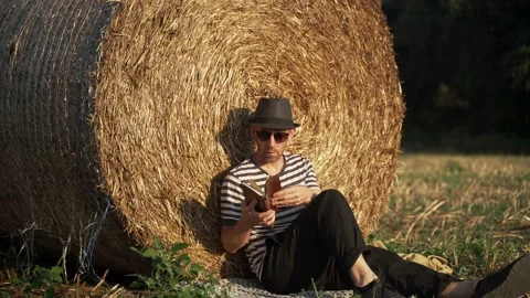 A young man chill in the field. A man in a stripe shirt and a black hat sitting Stock Footage 134459818