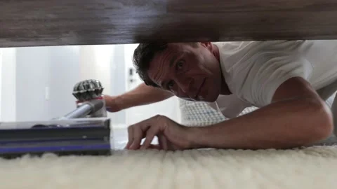 Young man cleaning a floor under a bed lying on the floor using vacuum cleaner Stock Footage 120812451