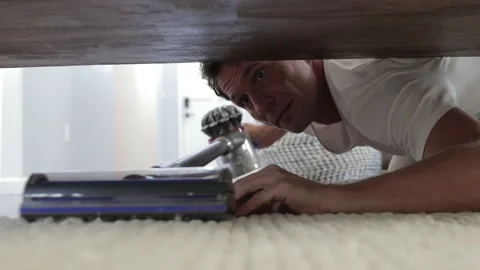 Young man cleaning a floor under a bed lying on the floor using vacuum cleaner Stock Footage 120812457