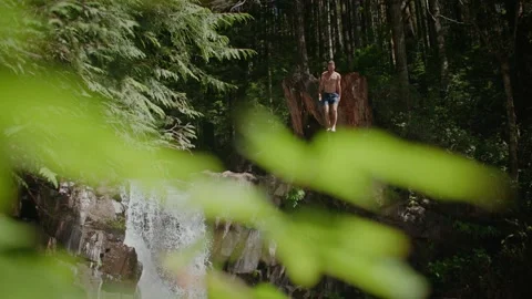 Young man cliff jumps next to waterfall in beautiful forest 库存影片 220301778