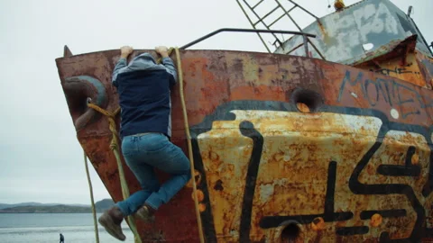 A young man climbs aboard an old rusty ship and stands on its edge Stock Footage 247285833