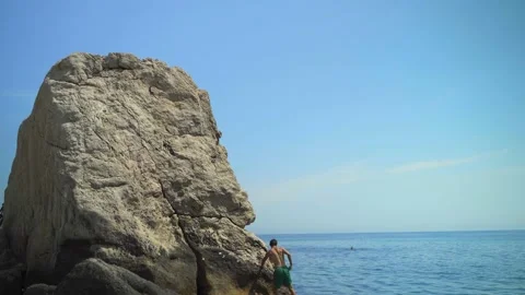 Young man climbs a high rock into the sea on a hot summer day to jump into the Stock Footage 157501233