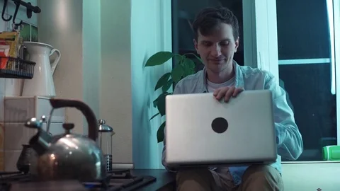 Young man closes computer sitting at kitchen while kettle boiling on stove Stock Footage 83871296