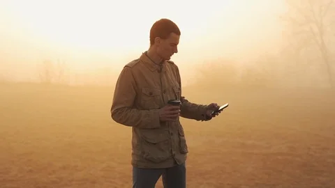 Young man in coat drinking his morning coffee and uses and scrolls mobile phone 库存影片 112586314