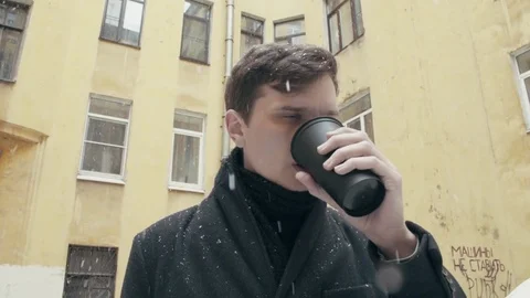 Young man in coat standing under snowfall drinking coffee from black plastic cup Video stock 74895084