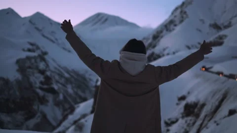 Young man in a coat on top of mountain summit at sunset, raises arms into air Stock Footage 169659898