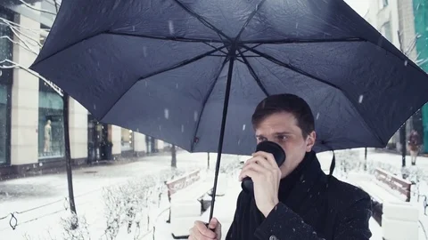 Young man in coat with umbrella under snowfall using smartphone on alley Stock Footage 74895401