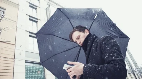 Young man in coat with umbrella under snowfall using smartphone on alley Stock-Footage 74895959