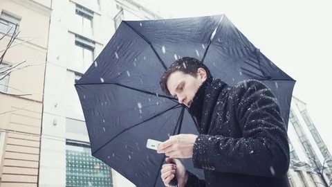 Young man in coat with umbrella under snowfall using smartphone on alley Video stock 74896093