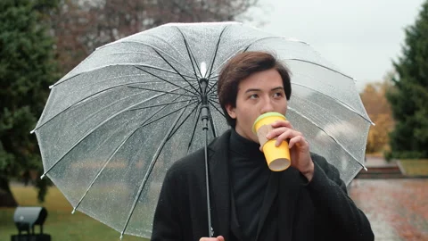 Young man in a coat walking under a transparent umbrella in the autumn city park Stock Footage 256695214