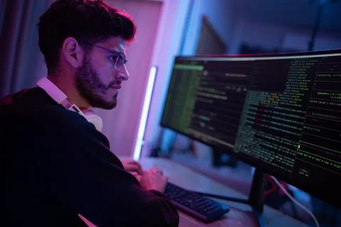 Young man coding on dual monitors in a dimly lit workspace during the night Stock Photos