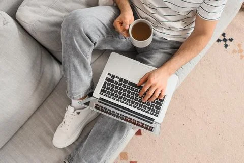 Young man with coffee using laptop to search online on sofa at home, top view Stock Photos