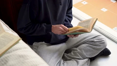 Young man in comfortable clothes reading an old book next to a window in a br Stock Footage 167089525
