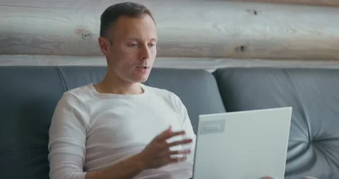 Young man communicates via video link using laptop while sitting on the sofa in Stock Footage 205575852