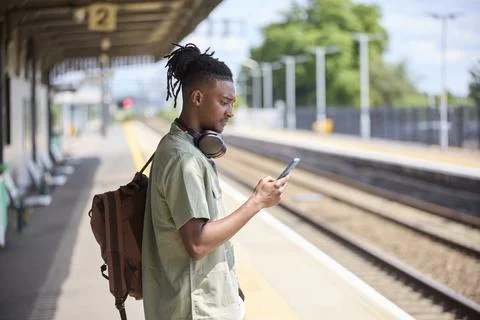 Young Man Commuting To Work On Train Standing  On Platform Looking At Mobil.. Stock Photos