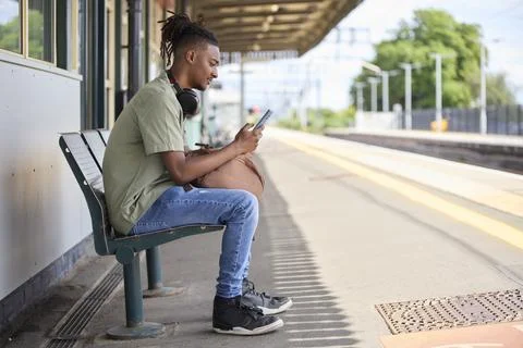 Young Man Commuting To Work On Train Sitting  On Platform Looking At Mobile.. Foto stock