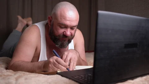 A young man with a computer on the bed lies on his stomach and makes notes in a Video stock 146662011