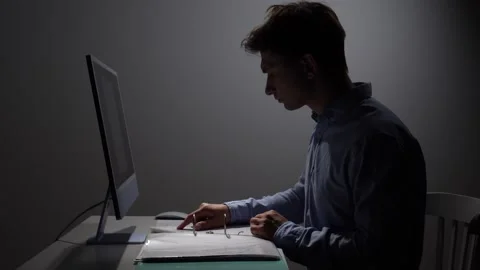 Young man concentrating on documentation and computer work in a dimly lit Stock Footage 342672466