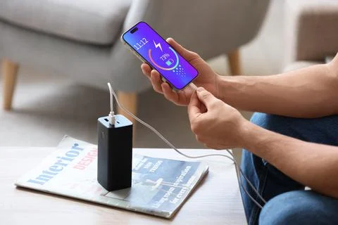Young man connecting charging cable of mobile phone to modern power bank at h Stock Photos