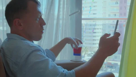 Young man contemplating at a large window with a cold drink and phone in hand Stock Footage 303816884