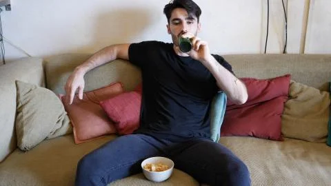 Young man on couch drinking beer and eating Stock Photos