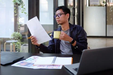 Young man creative designer holding coffee cup and reading documents in moder Stock Photos