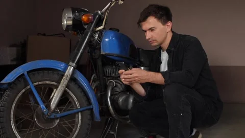 Young man crouching next to a vintage blue motorcycle, actively searching for Stock Footage 342771262