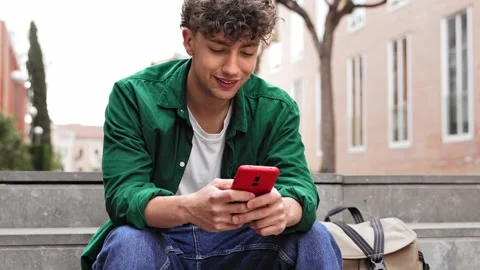 Young man with curly hair using red cell phone app in the city park Stock Footage 276735093
